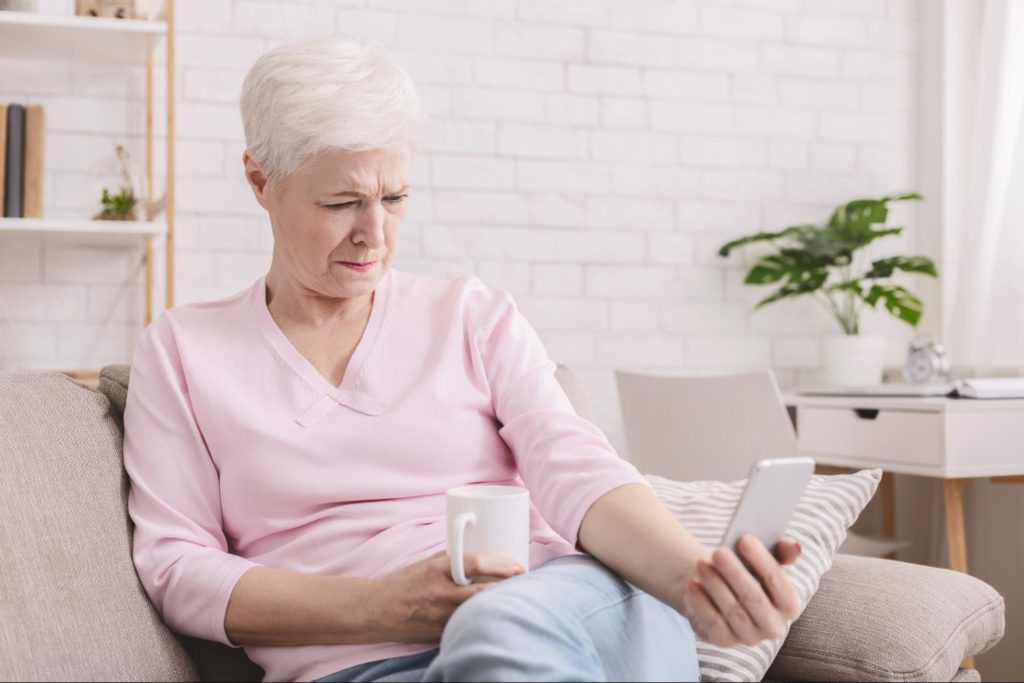 An older woman sitting on her sofa with a coffee cup struggling to read her phone as she holds it out in an extended arm.